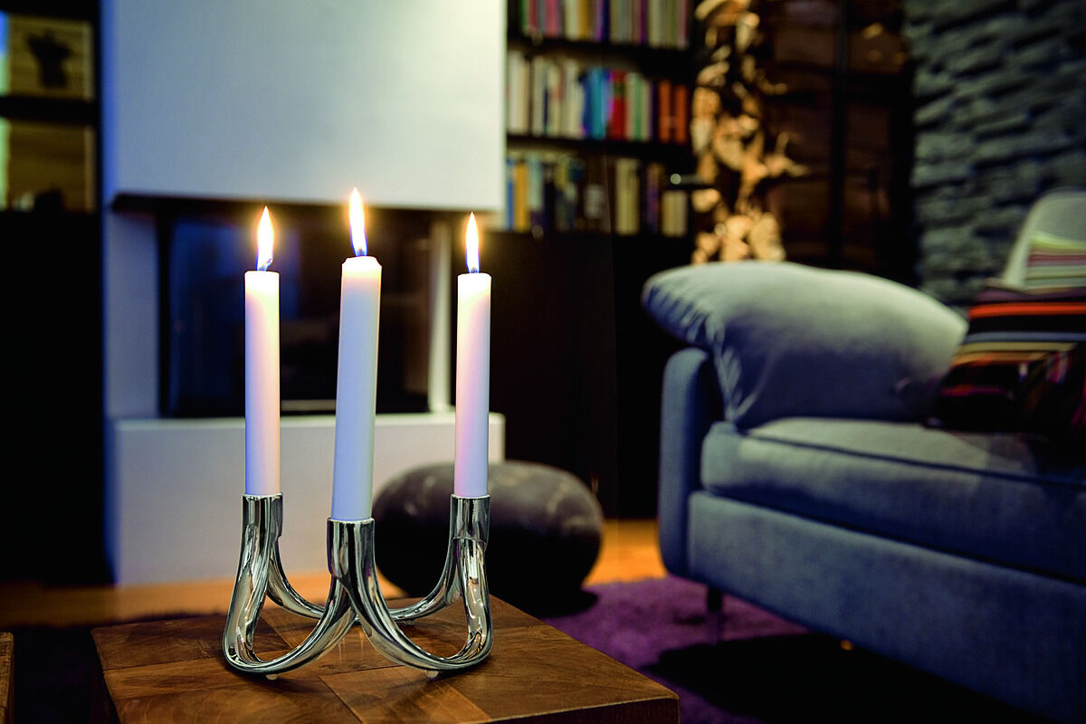 Das Bild zeigt einen eleganten magnetischen Kerzenhalter namens »BOW« mit drei brennenden weißen Kerzen. Der Kerzenhalter befindet sich auf einem Holztisch in einem stilvoll eingerichteten Wohnzimmer mit einem Bücherregal und einem grauen Sessel im Hintergrund, was eine gemütliche Atmosphäre schafft.