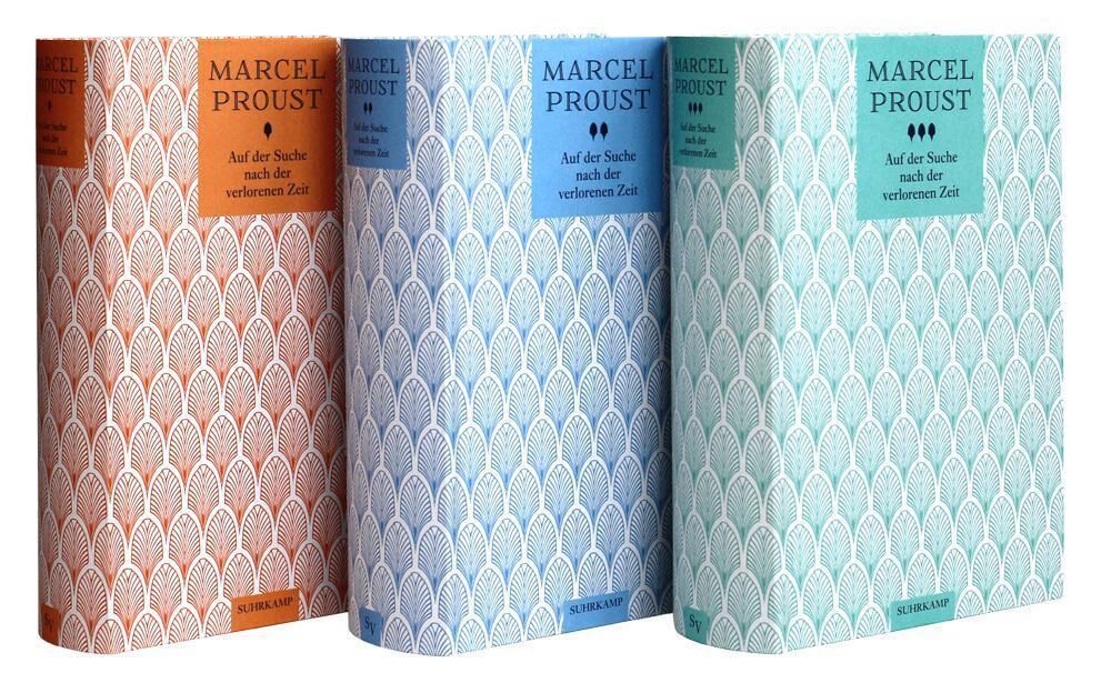 Auf dem Bild sind drei Bücher von Marcel Proust mit dem Titel 'Auf der Suche nach der verlorenen Zeit' zu sehen. Die Buchrücken sind in den Farben Orange, Blau und Türkis gehalten und weisen ein dekoratives Muster auf.