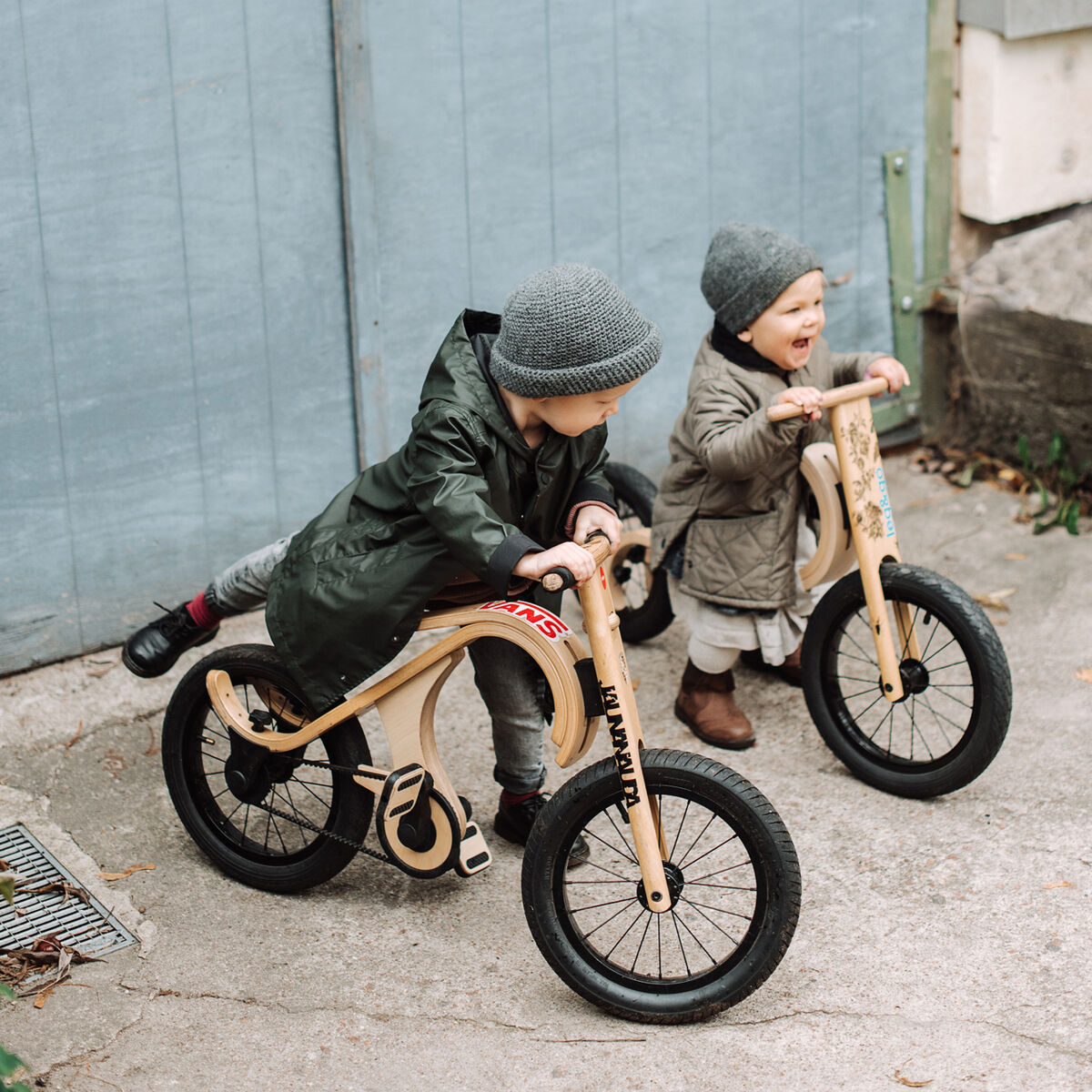 Zwei kleine Kinder spielen draußen auf Holzbikes mit schwarzen Reifen vor einem hellblauen Holztor. Beide Kinder tragen Mützen und Mäntel. Ein Kind hat die Füße auf Pedalen, das andere hebt den Fuß in Richtung Pedal. Die Szene vermittelt spielerisches Lernen, Gleichgewichtstraining und Bewegungsfreude.