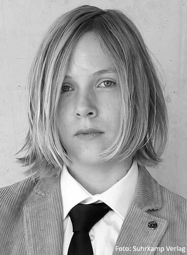 Schwarz-weiß Porträt eines jungen Menschen mit schulterlangem Haar, Anzug und Krawatte. Aufnahme von Suhrkamp Verlag.