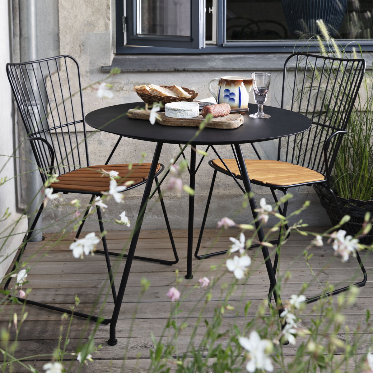 Zwei schwarze Esszimmerstühle PAON mit Holzlatten auf der Sitzfläche stehen an einem runden, schwarzen Tisch auf einer Terrasse. Im Hintergrund sind große Fenster und eine Wand zu sehen. Auf dem Tisch befinden sich verschiedene Leckereien und dekorative Elemente. Weiße Blumen ragen im Vordergrund in das Bild hinein.