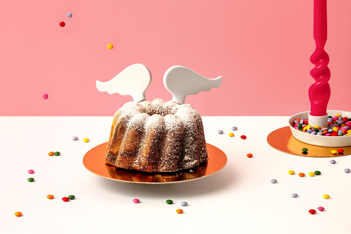 Ein Gugelhupfkuchen mit weißen Porzellanflügeln darauf, umgeben von bunten Zuckerstreuseln gegen einen rosa Hintergrund.