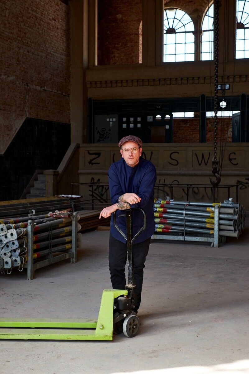 Ein Mann mit einer blauen Jacke und einer Kappe lehnt auf einem Hubwagen in einer Werkstatt. Im Hintergrund sind Metallrohre, ein großes Fenster mit Gitter mustern und grafitierten Wänden zu sehen.