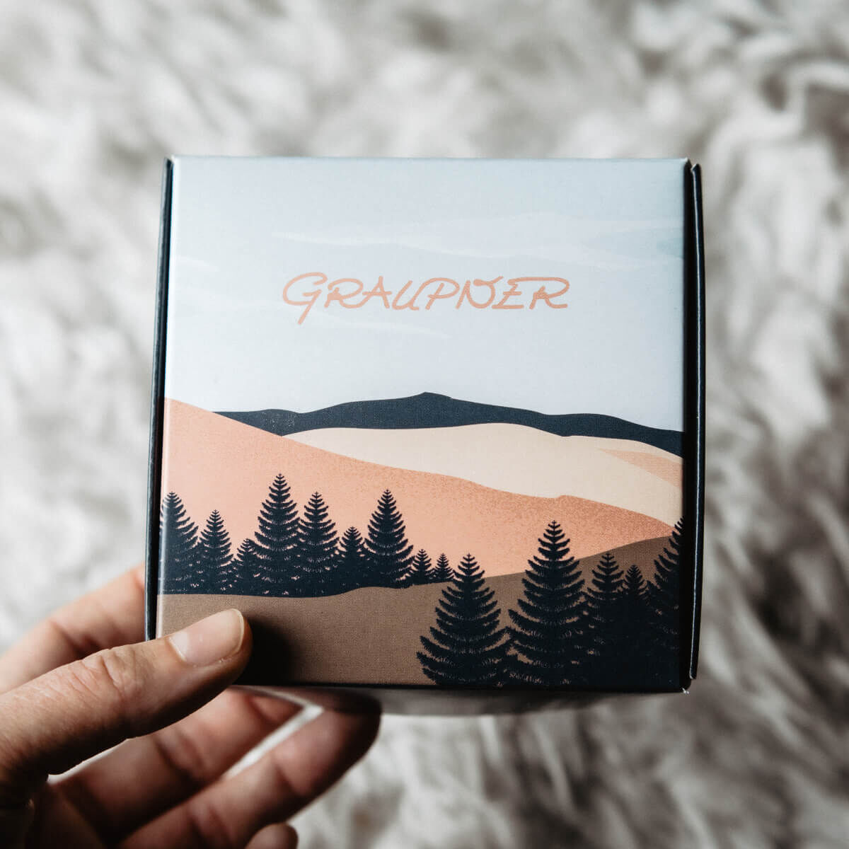 Eine Hand hält eine dekorative Verpackung mit der Aufschrift 'Graupner'. Die Illustration auf der Verpackung zeigt eine hügelige Landschaft mit Tannenbäumen in Brauntönen.