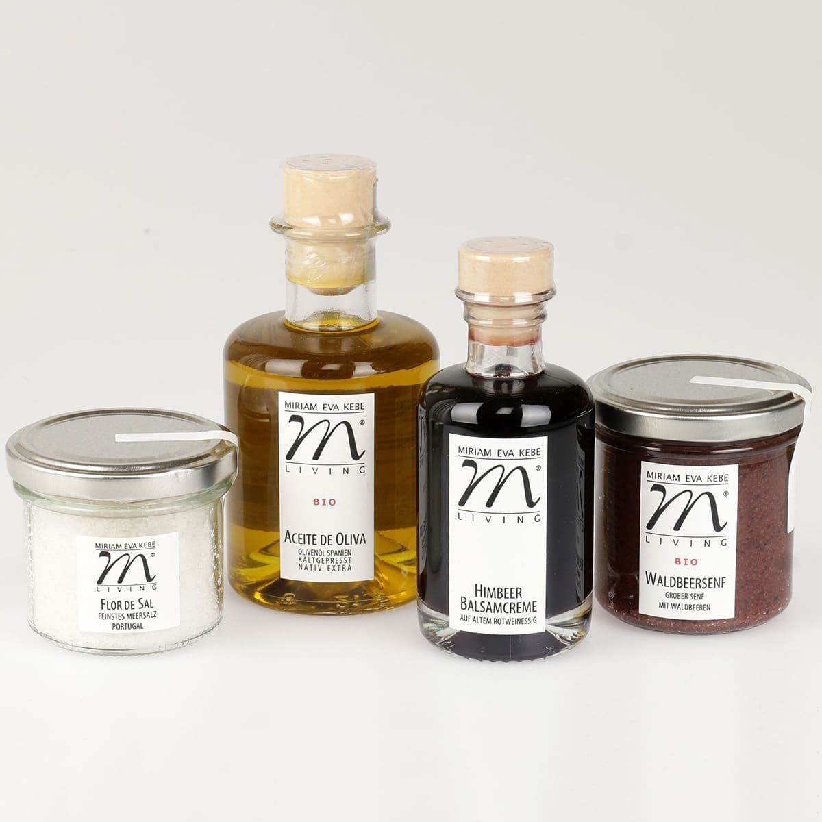 Ein Jubiläums-Set mit verschiedenen bio-zertifizierten Produkten: Fleur de Sal, Olivenöl, Himbeer Balsamcreme und Waldbeersensenf von Miriam Eva Kebe. Die Produkte sind in transparenten Gläsern und Flaschen verpackt, die jeweils mit einem Naturkorken verschlossen sind. Die Etiketten sind weiß und beinhalten schwarze Schrift sowie einen goldenen Logo-Akzent.