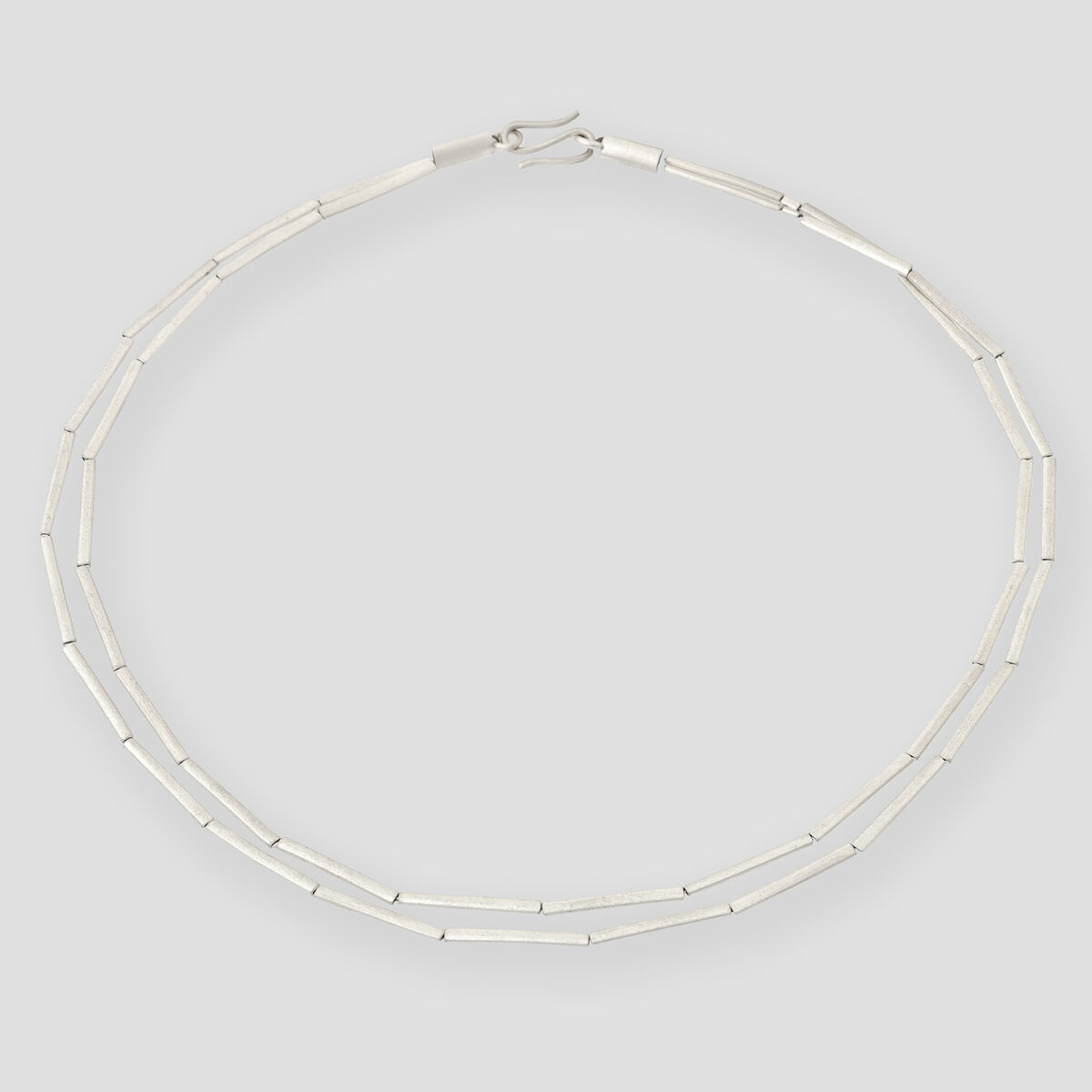 Das Bild zeigt ein elegantes Collier in Form einer Halmkette aus Silber, bestehend aus feinen, aneinandergereihten Stäben. Die Kette liegt kreisförmig auf einem hellgrauen Hintergrund.