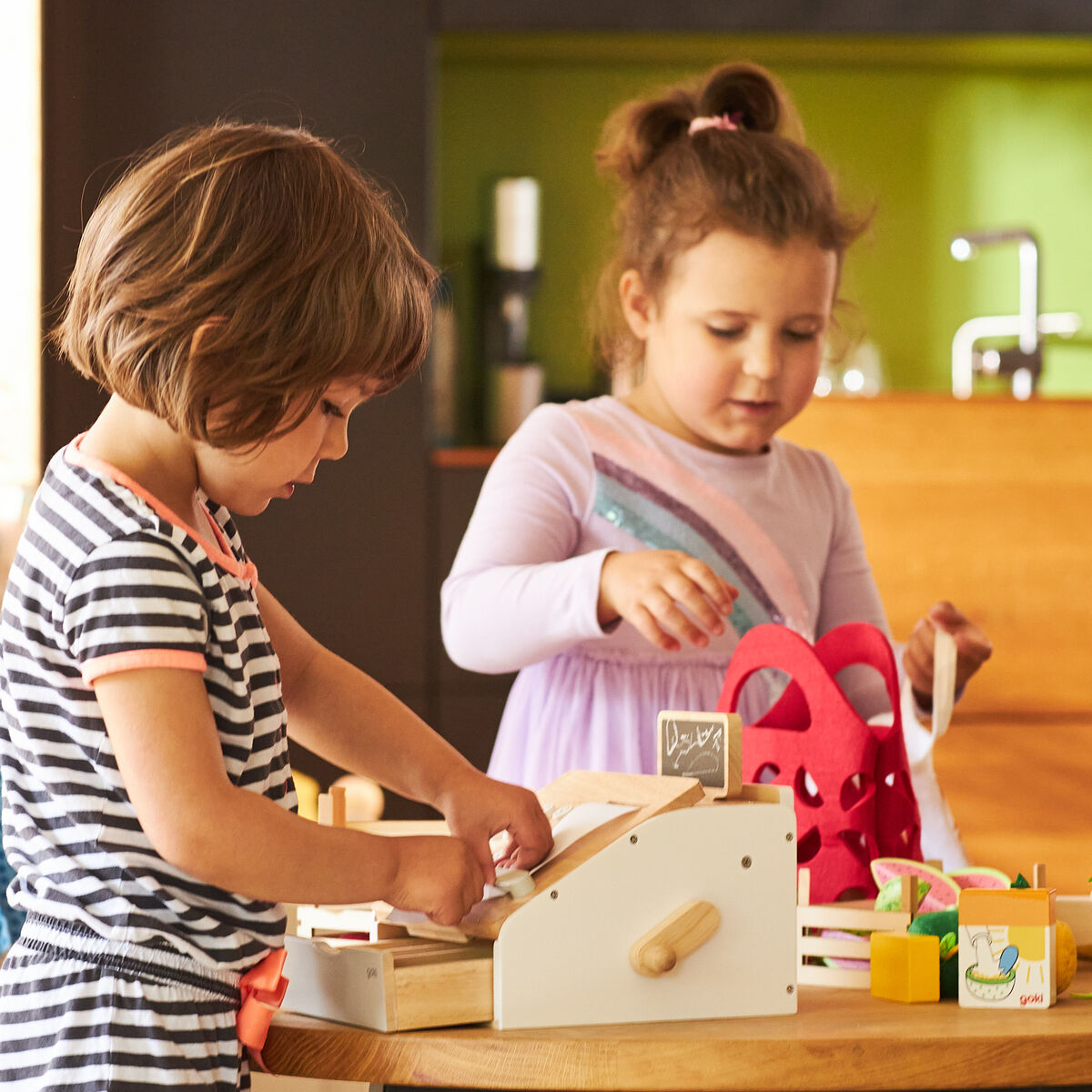 Zwei Kinder spielen gemeinsam mit einer Kaufmannsladenkasse aus Holz an einem Tisch. Verschiedene bunte Zubehörteile und Spielwaren sind um die Kasse verteilt. Die Szene findet in einer Wohnküche mit freundlicher Atmosphäre statt.