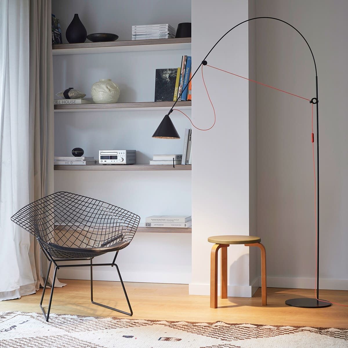 Eine moderne Stehlampe »AYNO« in einem minimalistischen Wohnzimmer mit einem schwarzen Schwenkarm und einem schwarzen Drahtstuhl vor einem weißen Bücherregal. Ein Holzhocker steht neben der Lampe, die auf einem hellen Holzboden platziert ist.