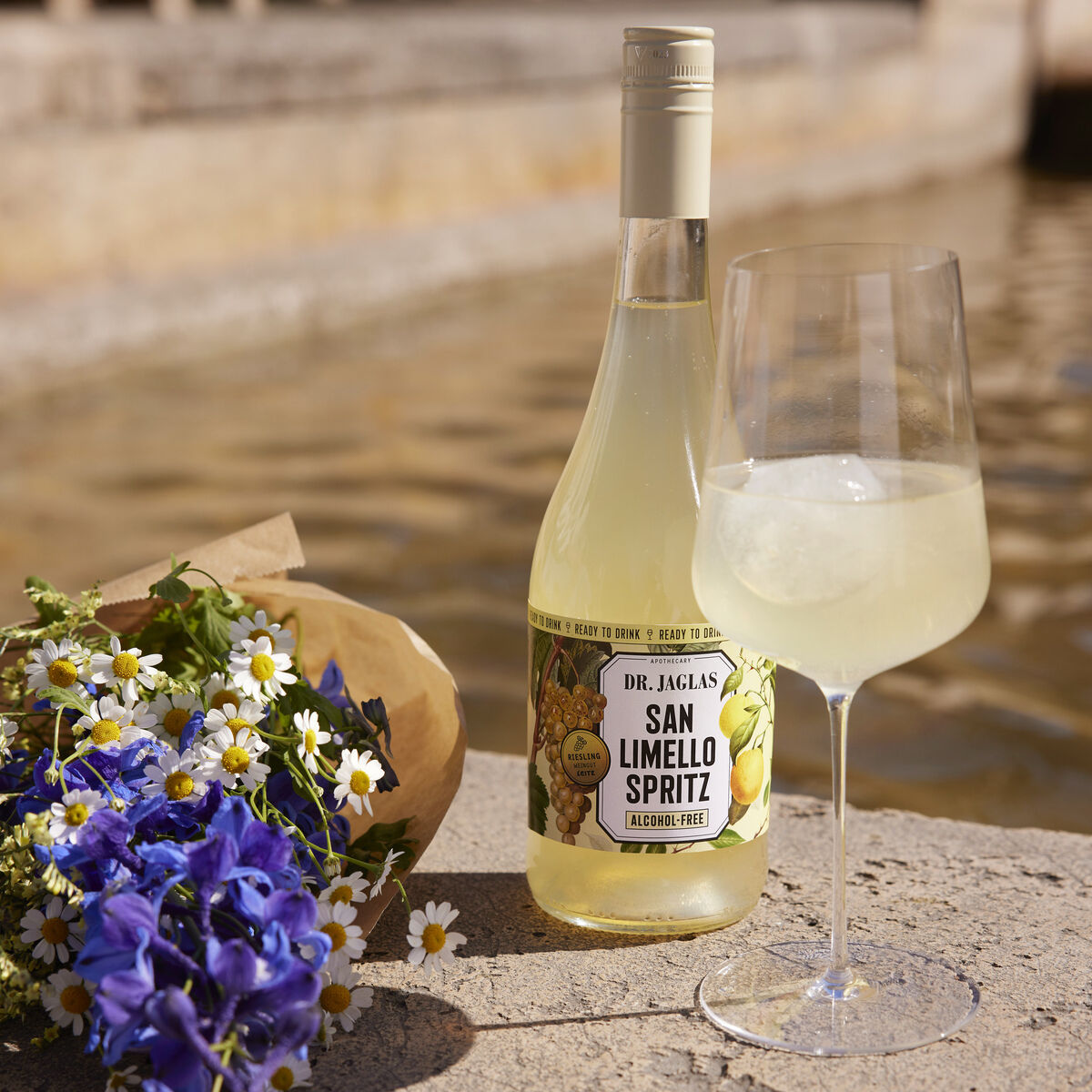 Eine Flasche San Limello Spritz steht auf einem Stein neben einem Glas mit dem Getränk und einem Blumenstrauß. Der Hintergrund zeigt einen verschwommenen Außenbereich mit Wasser.