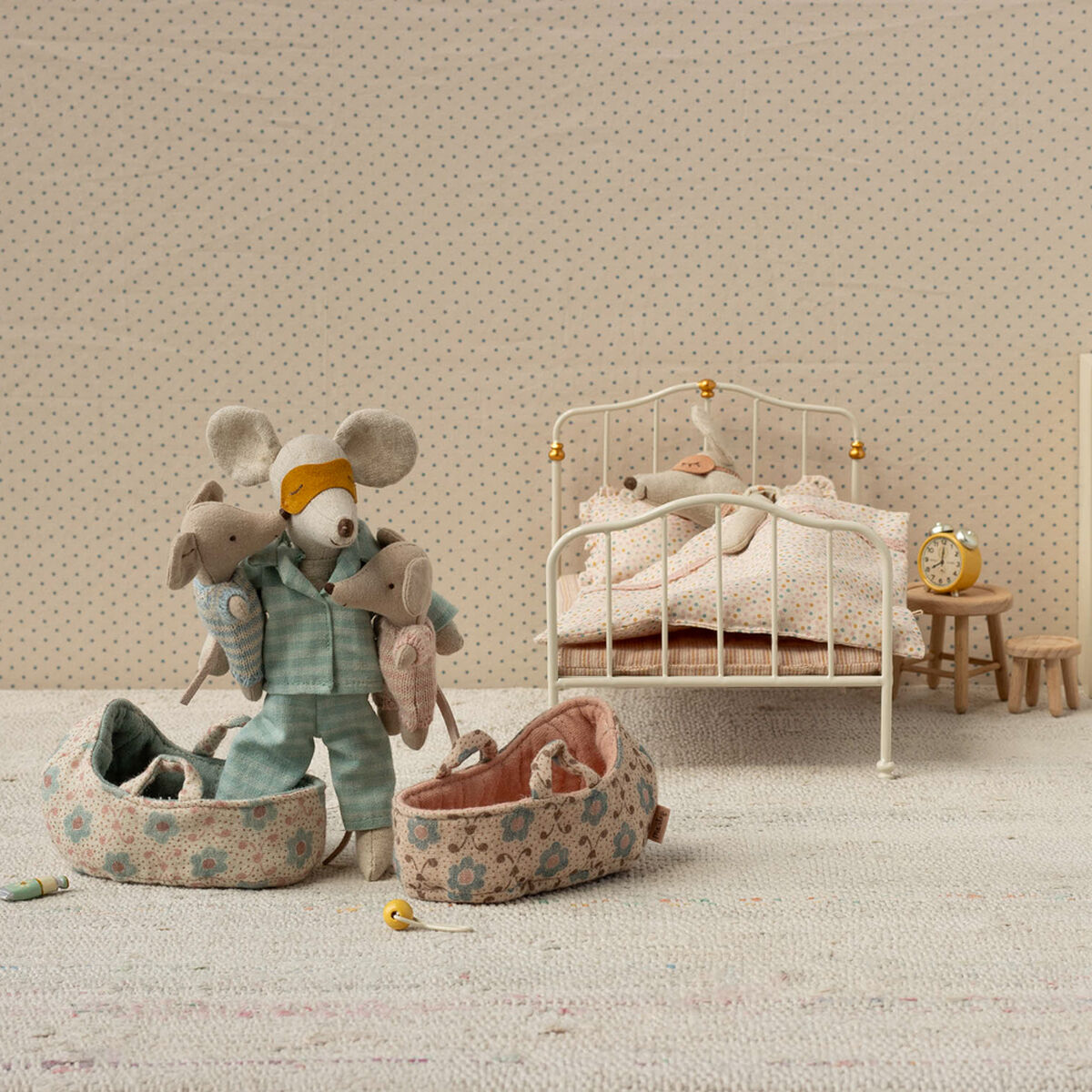 Eine kleine Stoffmaus liegt in einer rosafarbenen Tragetasche auf einem hellen Teppich in einem liebevoll gestalteten Kinderzimmer. Im Hintergrund sind ein kleines Bett, eine weitere Maus und verschiedene Möbelstücke zu sehen.