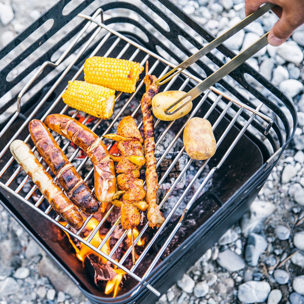 Ein Grillrost der Marke »CUBE« ist mit verschiedenen Lebensmitteln wie Würsten, Maiskolben und einer Kartoffel über einer Flamme belegt. Eine Hand hält Grillzangen, um das Grillgut umzudrehen. Die Szene findet auf einem steinigen Untergrund statt.