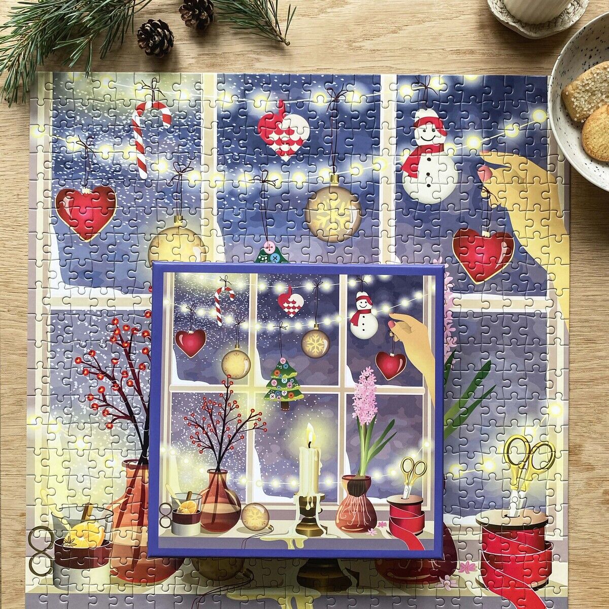 Das 500 Teile Puzzle »Weihnachtsfenster« von ViSSEVASSE zeigt ein festlich dekoriertes Fenster mit Schneemännern, roten Herzen, goldenen Kugeln, Kerze und Tannengirlande. Im Vordergrund sorgen Deko, Keksteller, Tannenzweig und Kerze für eine warme, gemütliche Atmosphäre.