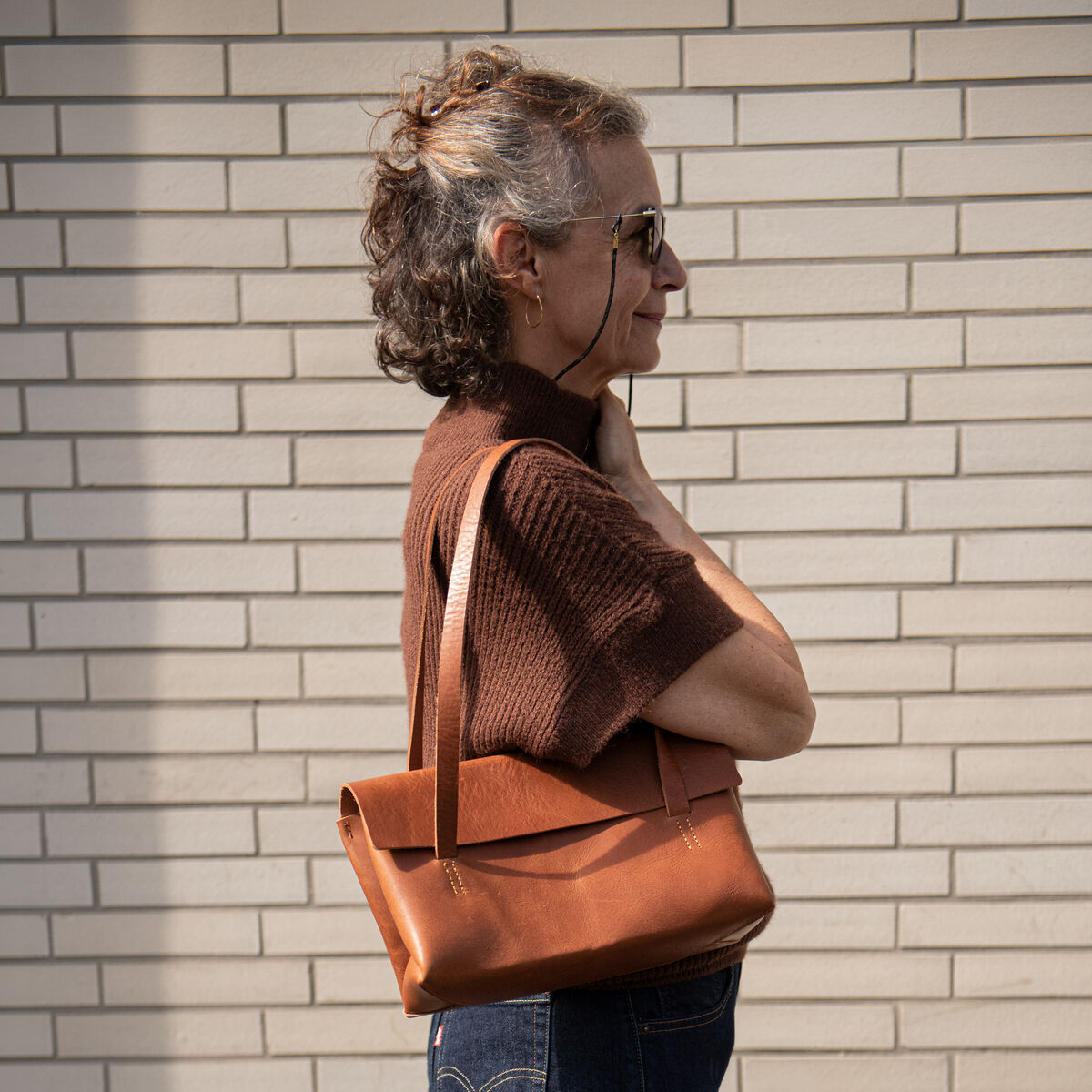 Seitliche Ansicht einer Frau, die eine cognacfarbene Kernleder Schultertasche von Harold's Lederwaren über der Schulter trägt. Sie steht vor einer hellen Backsteinwand, trägt braunen Pullover und dunkelblaue Jeans. Die Tasche wirkt hochwertig und elegant.
