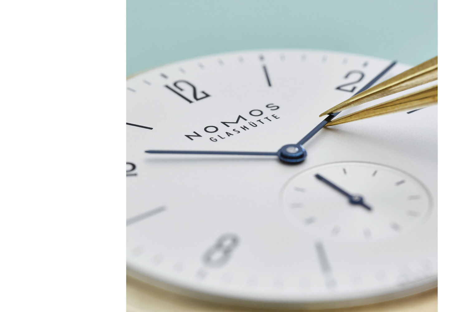 Detailaufnahme des Zifferblatts der NOMOS ZEIT-Sonderedition »Tangente Gold neomatik 38 Update«. Emailleblaues Zifferblatt, feine rhodinierte Zeiger, goldene Pinzette richtet Zeiger. Die Uhr zeigt minimalistisches Design und präzise Handwerkskunst.