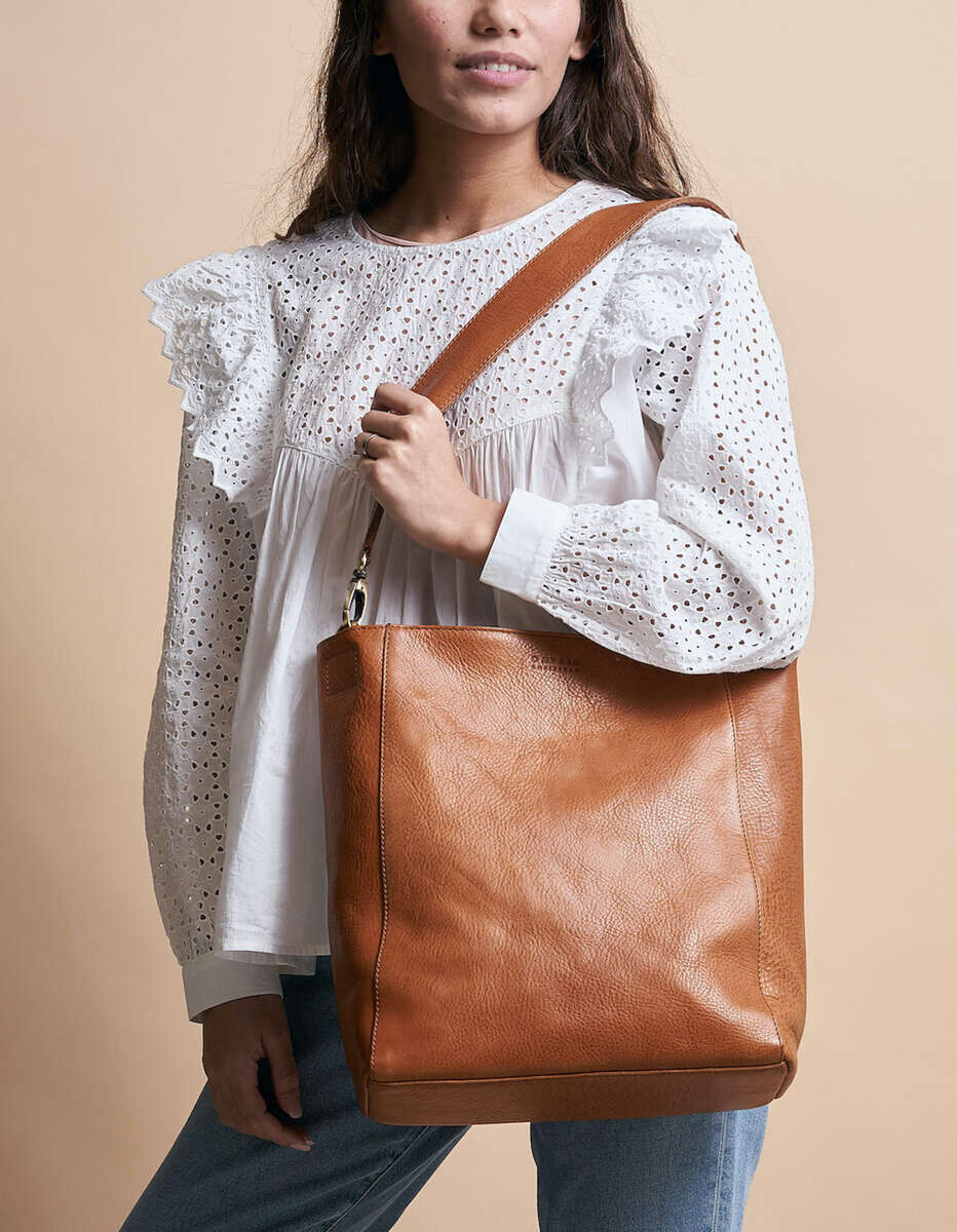 Eine Frau trägt eine braune Ledertasche »Sofia«, begleitet von einer weißen Spitzenbluse und blauen Jeans, vor einem beigen Hintergrund.