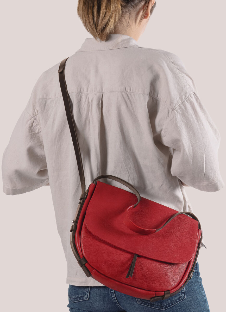 Eine Frau trägt eine rote Schultertasche »Slingbag M« mit einem verstellbaren braunen Gurt. Sie trägt eine weiße Bluse und eine blaue Jeans und ist von hinten zu sehen.