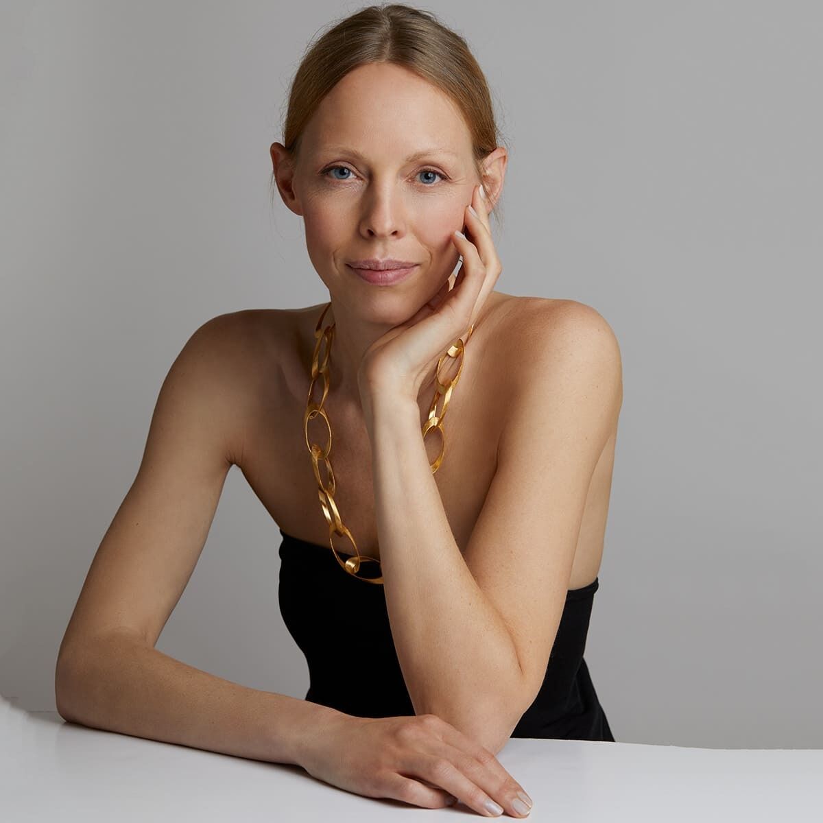 Eine Frau trägt eine elegante goldene Halskette. Sie hat langes blondes Haar und trägt ein schwarzes Oberteil. Der Hintergrund ist neutral grau.