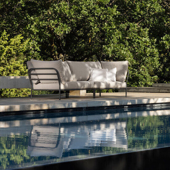 Eine Lounge-Eckcouch aus wetterfesten Materialien, die an einem sonnigen Tag auf einer Terrasse neben einem Pool steht, umgeben von viel Grün und minimalistischer Architektur.