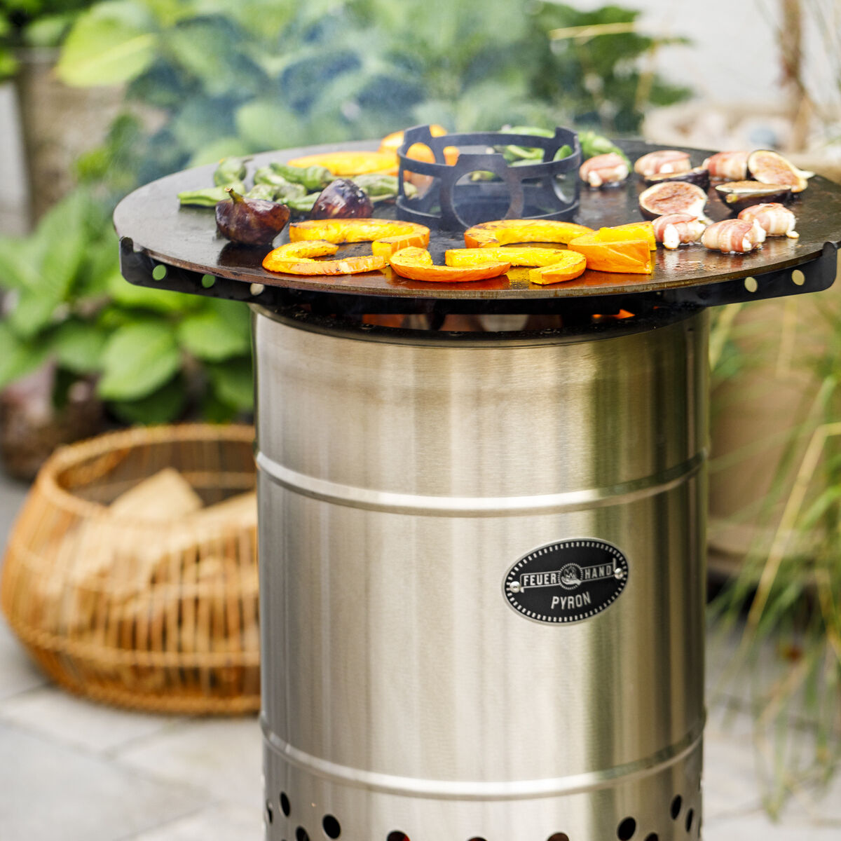 Eine »Pyron« Platte in einem Garten, wo auf der runden Edelstahlplatte verschiedene Gemüse und Fleischstücke gegrillt werden. Im Hintergrund sind Pflanzen und Gartenzubehör zu sehen.