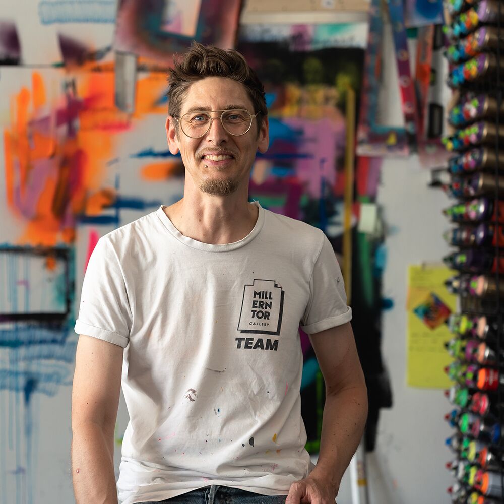Ein Mann steht in einem kreativen Atelier vor einem farbenfrohen, abstrakten Kunstwerk mit dominierenden Pink- und Orangetönen. Er trägt ein weißes T-Shirt mit der Aufschrift 'Millentor Gallery Team', eine Brille und lächelt freundlich in die Kamera. Im Hintergrund sind weitere bunte Kunstwerke, Farbe und Zeichenmaterialien zu sehen.