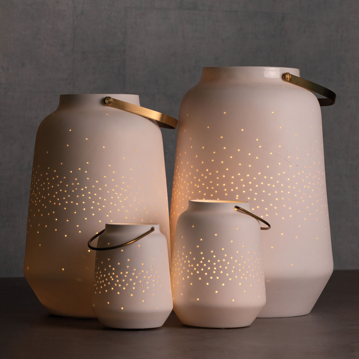 Vier weiße Porzellanlaternen unterschiedlicher Größe, die auf einem Tisch stehen und mit warmem, goldenen Licht leuchten. Die Laternen haben eine schlichte, moderne Form mit kleinen, glänzenden Löchern, durch die das Licht strahlt.