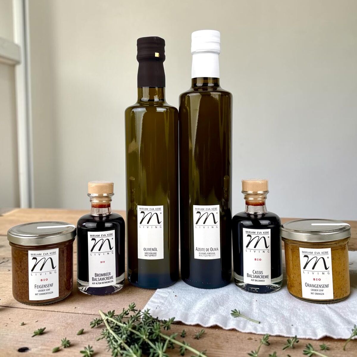 Sechs Feinkostprodukte aus der Kebe Living Manufaktur stehen auf einem Holzbrett: zwei Olivenölflaschen, zwei Balsamcremes in kleinen Flaschen und zwei Senfgläser. Davor frische Kräuter, Hintergrund hell und schlicht. Hochwertige Aufmachung.