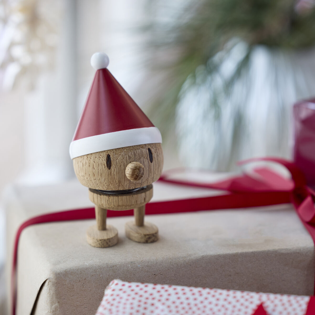 Die Holzfigur Hoptimist Elf Bumble Raw oak von Hoptimist steht mit roter Zipfelmütze auf einem Geschenk mit rot-weißem Band. Im Hintergrund weitere festlich verpackte Geschenke und unscharfe Dekoration. Weihnachtsatmosphäre vor einem Fenster.