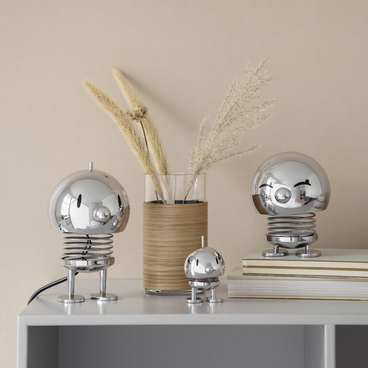 Ein Hoptimist Bumble Chrome auf einem Regal, umgeben von anderen dekorativen Gegenständen. Die Szene zeigt drei unterschiedlich große, glänzende, silberfarbene Hoptimist-Figuren auf einem weißen Regal, mit beigen und braunen Büchern sowie einem Vase mit getrockneten Gräsern im Hintergrund.