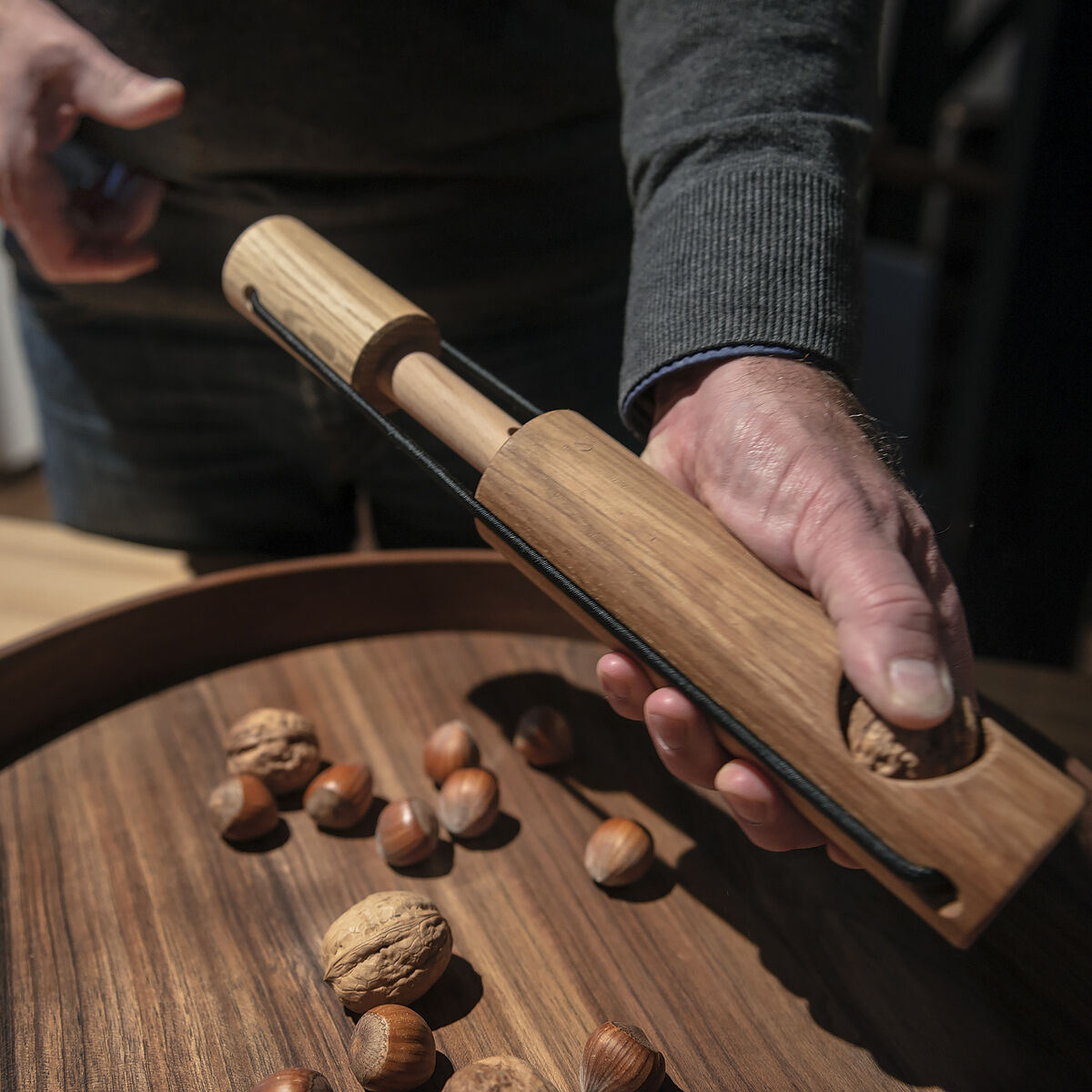 Ein hölzerner Nussknacker namens »Kracher« wird von einer Person benutzt, um Walnüsse zu knacken. Der Nussknacker liegt auf einer Holzschale zusammen mit Walnüssen und Haselnüssen; eine männliche Hand hält das Gerät. Verschiedene Brauntöne dominieren das Bild.