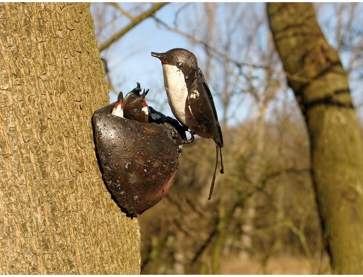 Ein künstliches Schwalbennest aus Metall ist an einem Baumstamm befestigt. Das Nest ist künstlerisch gestaltet und zeigt eine Schwalbe mit einem Jungvogel darin. Im Hintergrund befinden sich verschwommene Bäume und blauer Himmel.