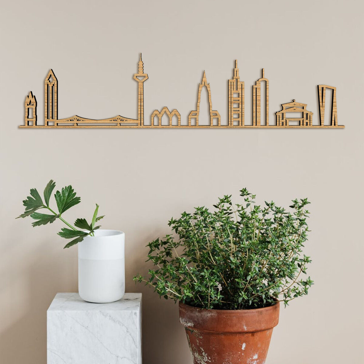 Holz-Silhouette von Frankfurts Skyline, aufgehängt an einer beigen Wand. Darunter steht ein Marmorpodest mit einer weißen Vase und einer grünen Pflanze sowie ein großer Tontopf mit einem größeren grünen Strauch.