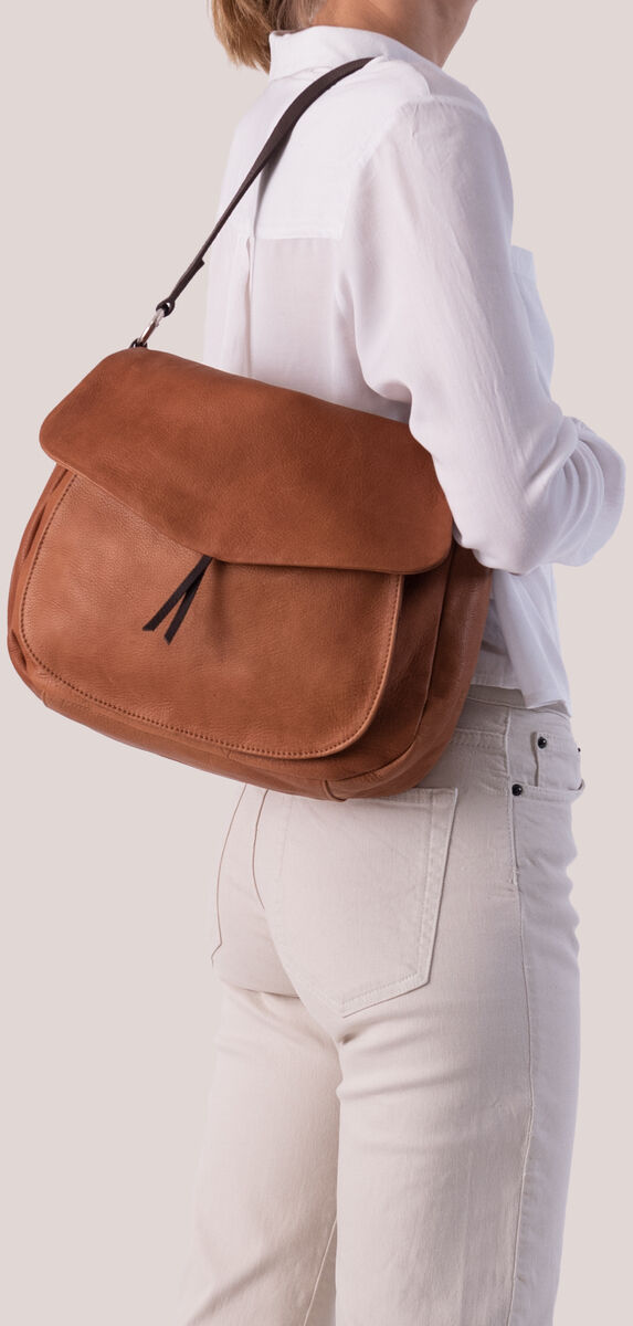 Eine Frau trägt eine braune Lederslingbag über ihrer Schulter. Sie trägt eine weiße Bluse und beige Hose. Die Tasche hat einen minimalistisch und stilvoll gestalteten Look.