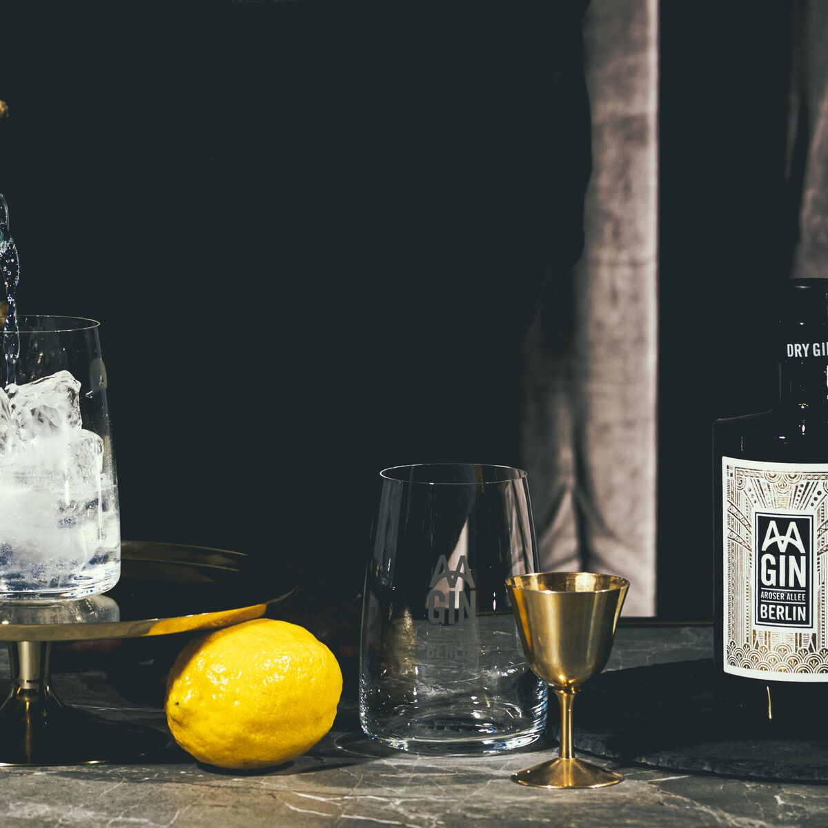 Ein Glas wird mit Gin Tonic befüllt und ist mit Eiswürfeln und Zitronenstücken garniert. Daneben steht eine geöffnete Flasche »AAGIN« Dry Gin, eine Zitrone und eine goldene Messbecher.