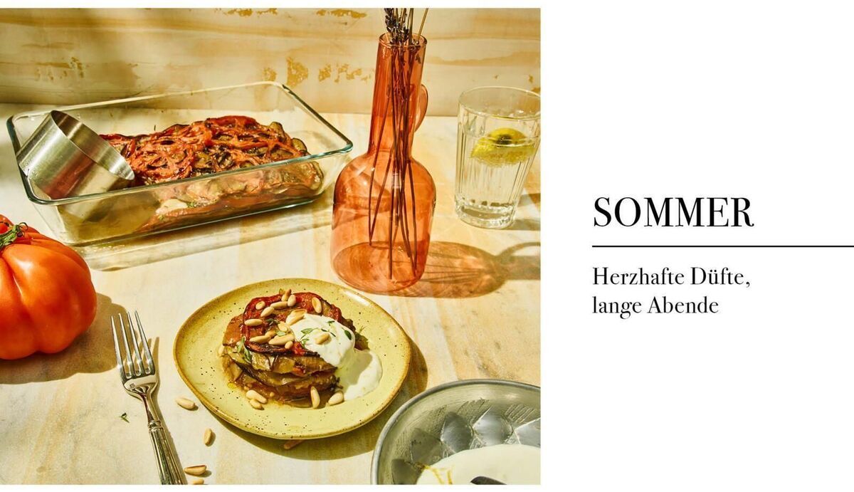 Auf einem hellen Tisch stehen ein Ofengericht in einer Auflaufform, eine gebratene Tomatenscheibe auf einem Teller mit Mandeln und Käse, eine orangefarbene Vase, ein Glas mit Zitronenwasser und eine große frische Tomate. Text: "SOMMER – Herzhafte Düfte, lange Abende".