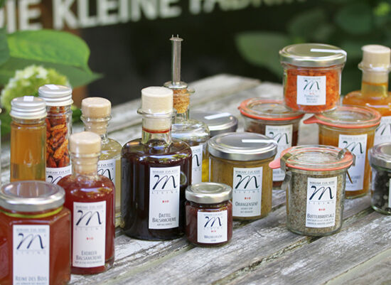 Verschiedene Gläser und Flaschen mit Gewürzen, Ölen und Marmeladen auf einem Holztisch, Etiketten mit Buchstabe 'M'
