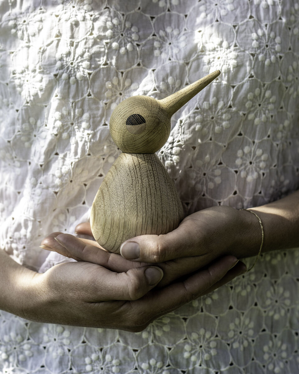 Zwei Hände halten eine aus Holz geschnitzte Vogelfigur namens „Large Natural Birds“. Die Figur hat eine natürliche Holzfarbe und ist minimalistisch gestaltet. Der Hintergrund zeigt ein helles, florales Muster.