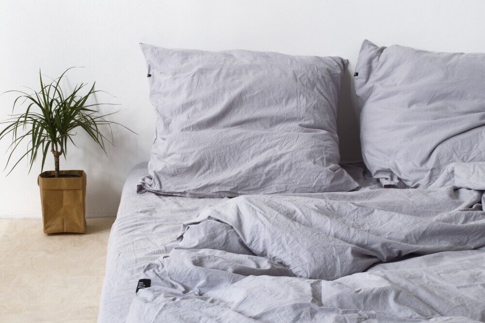 Eine gemütlich aussehende Bettwäsche in grauer Farbe auf einem Bett, daneben eine grüne Zimmerpflanze in einem braunen Topf. Die Wand im Hintergrund ist weiß.