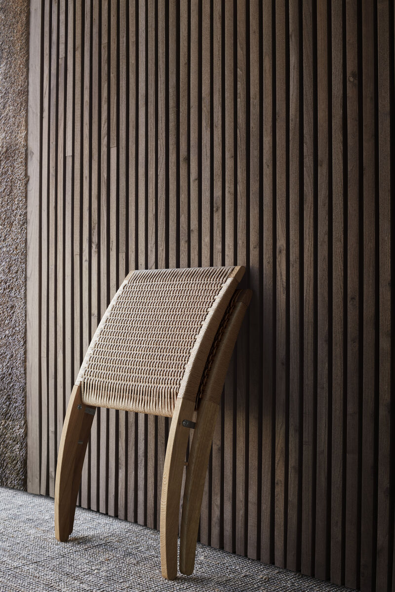 Der MG501 Cuba Chair ist ein eleganter Holzstuhl mit einer gewebten Sitzfläche und geschwungenen Linien, der vor einer vertikalen, braunen Holzplankenwand steht.