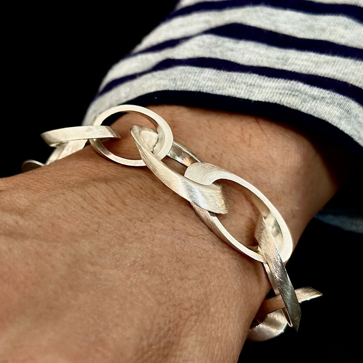 Ein silbernes Armband namens "Ellipse", getragen am Handgelenk einer Person, auf schwarzem Hintergrund, mit weiß gestreiftem Ärmel.