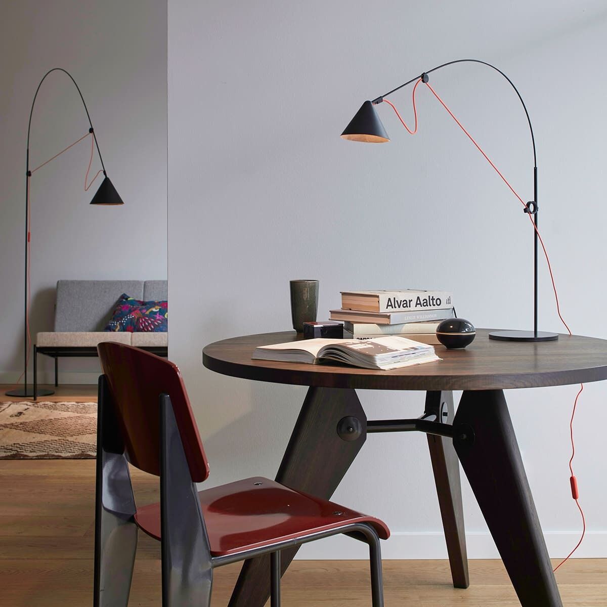 Moderne, minimalistische Stehlampe AYNO mit schwarzem Lampenschirm und rotem Kabel in einem stilvoll eingerichteten Raum. Die Lampe steht neben einem runden Holztisch mit Büchern, Tassen und Stühlen.