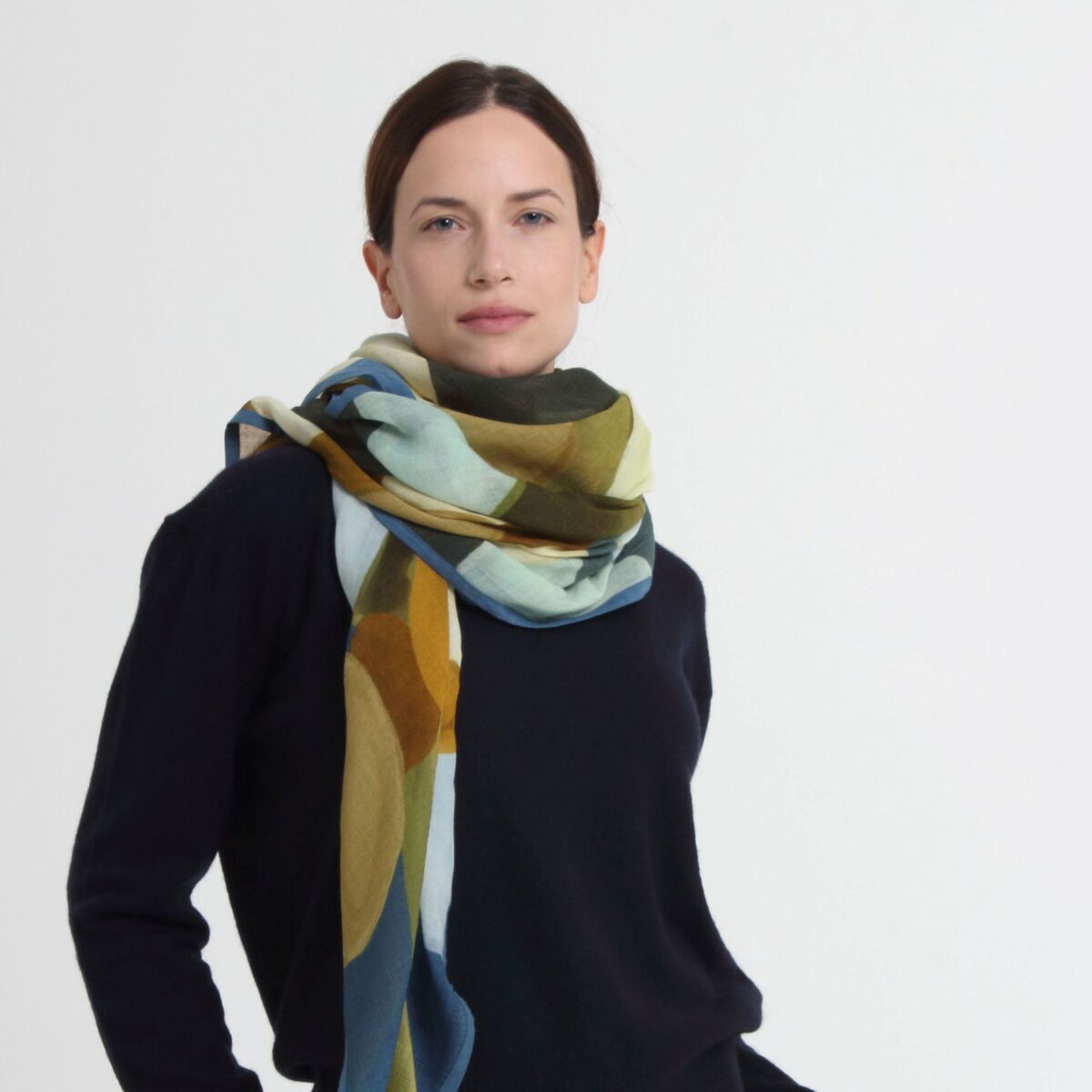 Eine Frau trägt einen Wollschal »Evie« Blau-Camel von Shalls Barcelona über einem schwarzen Pullover und weißen Hosen. Der Schal zeigt ein auffälliges, geometrisches Muster in Blau-, Grün-, Camel- und Gelbtönen. Der Hintergrund ist neutral und hell.