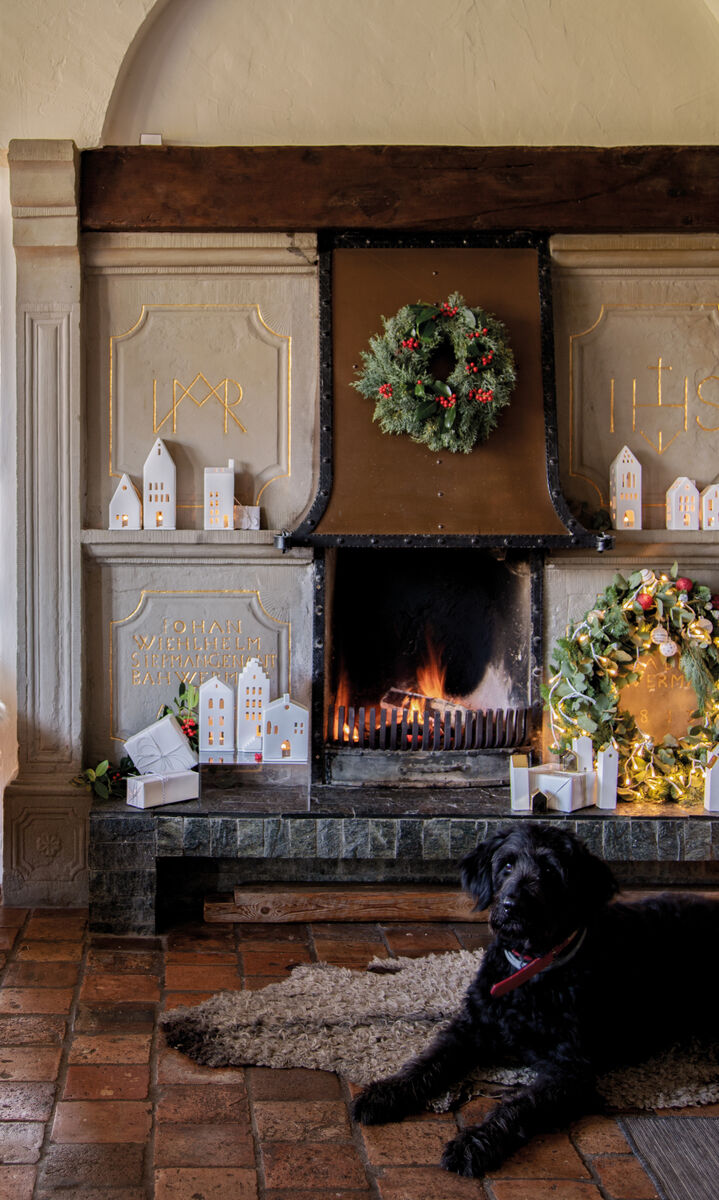 Gemütliches Wohnzimmerdekor mit einem Kamin, der von weihnachtlichen Lichthäusern, Kränzen und anderen festlichen Dekorationen umgeben ist. Ein schwarzer Hund liegt vor dem Kamin auf einem Teppich.
