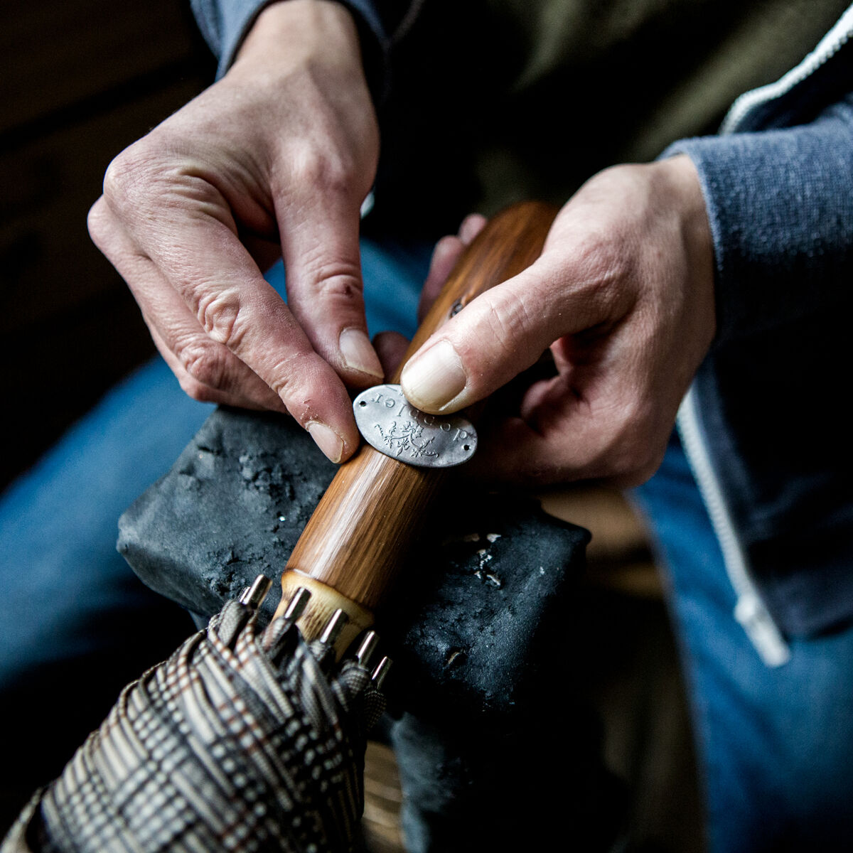 Eine Nahaufnahme von Händen, die an einem Regenschirm vom Modell »Norfolk Oxford« in der Farbe ‚Berry‘ arbeiten. Der Schirm hat einen Holzgriff und ein gemustertes Material. Es handelt sich um eine manuelle Reparatur oder Fertigung, die handwerkliches Können zeigt.
