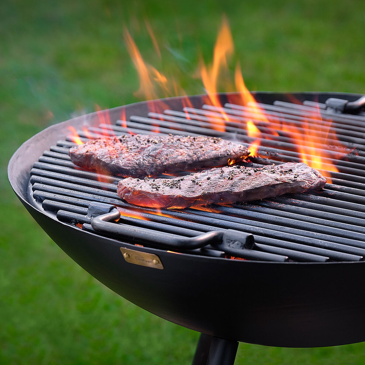 Runder Grillrost mit brennenden Flammen und zwei Steaks, die auf dem Grillrost garen. Scharfer Rasen im Hintergund.