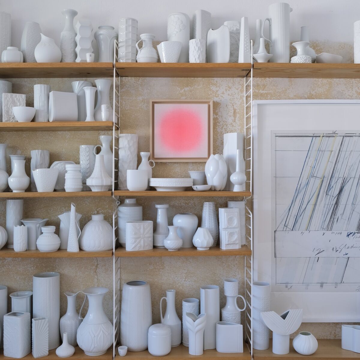 Ein modernes Wohnzimmerregal mit zahlreichen weißen Keramikvasen und Skulpturen, zentral hebt sich die ZEIT-Sonderedition Sorbet »Rot« als quadratisches Kunstwerk mit leuchtendem Rotton von der Wand ab. Daneben hängt ein großes, abstraktes Bild. Die Regalböden und Wand wirken hell und minimalistisch.