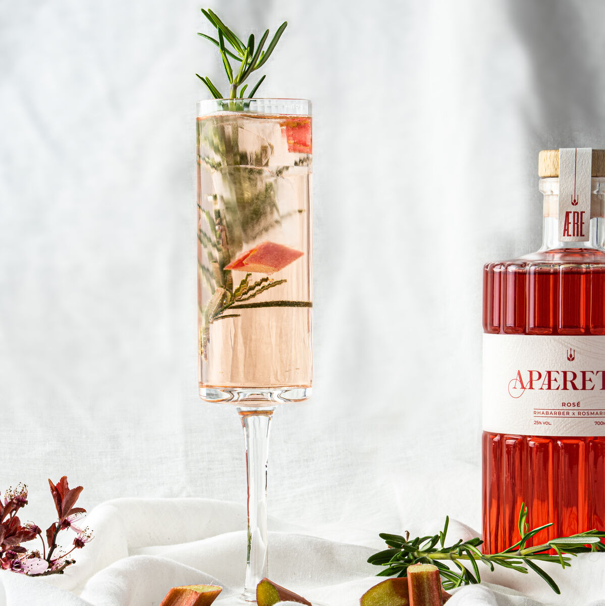 Ein Glas AERE Aperitif »Rosé« steht auf einem weißen Leinentuch, umgeben von Rosmarinzweigen und Rhabarberstücken. Im Hintergrund ist eine dazugehörige Flasche des Aperitifs zu sehen.