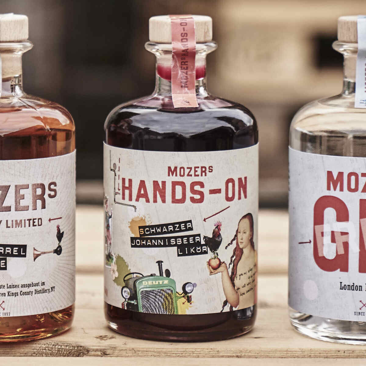 Fünf Flaschen Spiritsuosen der Marke Mazer Mozer auf einem Holztisch, einschließlich Johannisbeerlikör, Gin, und weiterer handgemachter Produkte, vor einem rustikalen Hintergrund aus Holzpaletten.