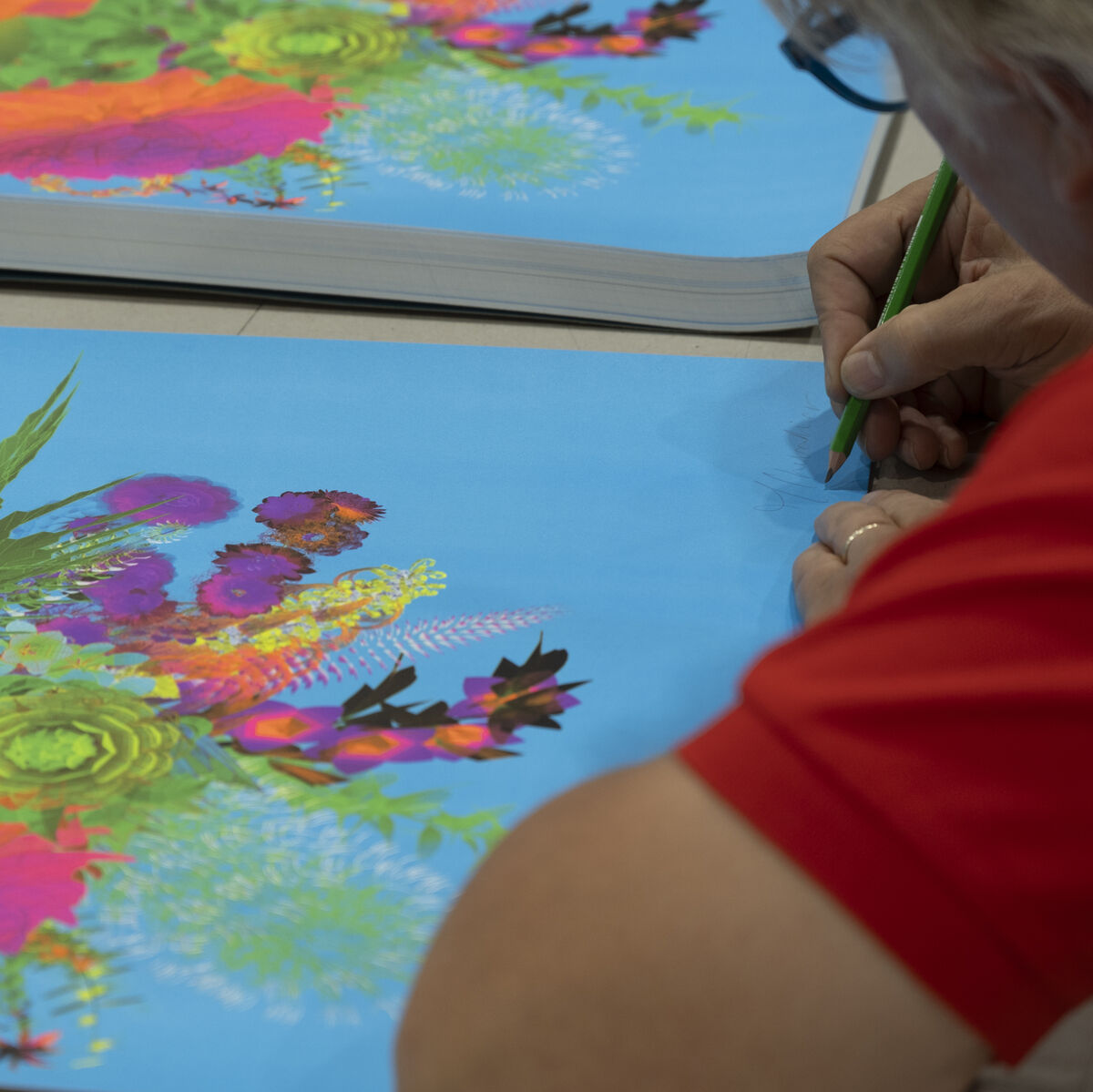 Künstler arbeitet an lebhaftem, farbenfrohen Kunstwerk auf Leinwand mit floralen und abstrakten Mustern in Blau-, Grün-, Orange-, Pink- und Lilatönen. Die Szene zeigt den kreativen Arbeitsprozess der digitalen Kunst auf einem Tisch.