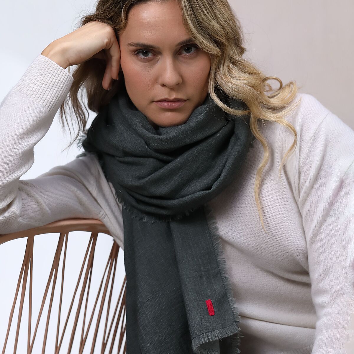 Frau mit einem doppelseitigen, grauen Wollschal »Carla«, sitzt in einem Stuhl und trägt einen hellen Pullover. Der Schal ist voluminös und elegant um den Hals geschlungen, kleiner roter Akzent am Etikett sichtbar. Moderne, ruhige Atmosphäre.
