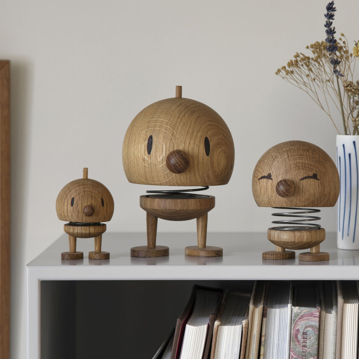 Drei Hoptimist Bumble Eiche Figuren in verschiedenen Größen stehen auf einem Regal vor einer hellen Wand. Die Figuren sind aus Holz gefertigt und haben eine minimalistische, skandinavische Optik. Neben den Figuren stehen ein Gefäß mit getrockneten Blumen und ein gerahmtes Bild.